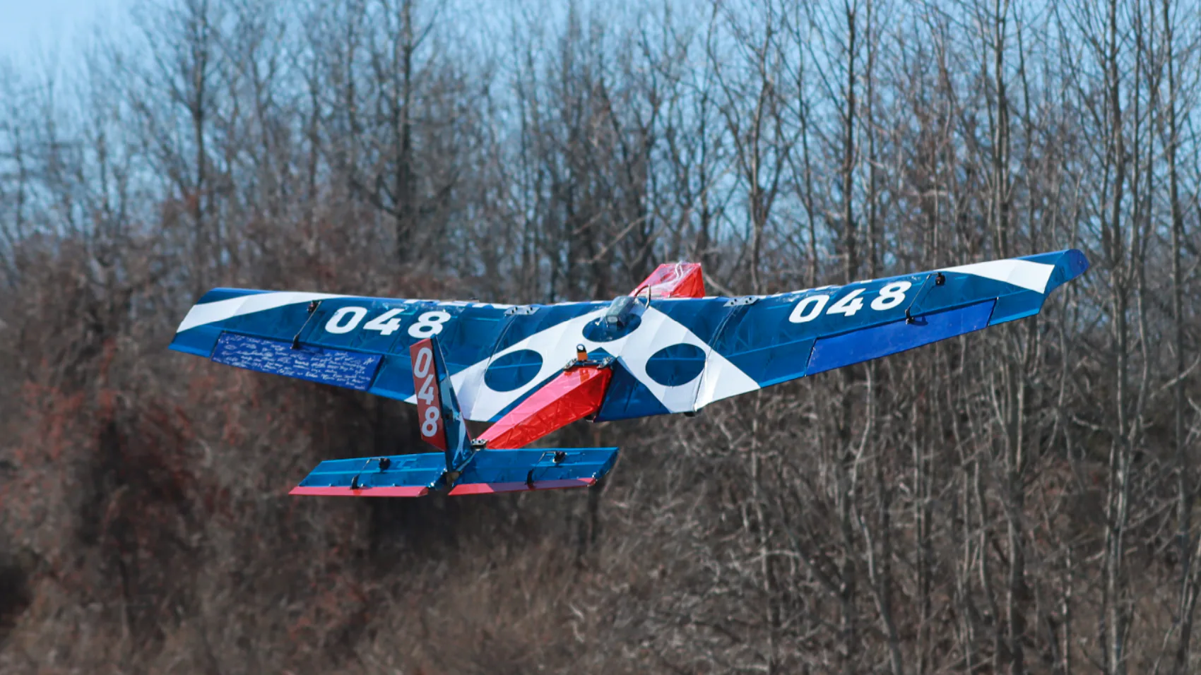 Rear view of the blue, white, and red Leviathan aircraft flying above a winter tree line.